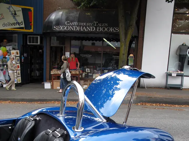 In this picture there is a blue color vehicle on the road and there are few stores and people in...